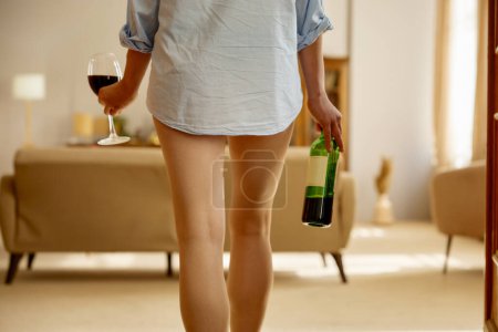 Téléchargez les photos : Jeune femme sensuelle portant une chemise masculine tenant une bouteille de vin et un verre à vin recadré tourné en mettant l'accent sur les jambes féminines minces - en image libre de droit