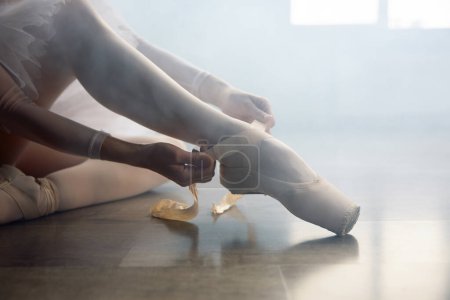 Téléchargez les photos : Ballerine assise sur le sol et portant des chaussures pointes avant la leçon de chorégraphie. Belle danseuse se préparant pour la performance ou l'entraînement à l'école de danse - en image libre de droit