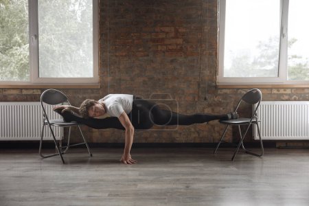 Foto de Flexible hombre bailarín de ballet haciendo estiramiento realizando split con dos sillas en estudio de danza moderna - Imagen libre de derechos