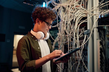 Photo for Serious young IT man wearing headphones working in data center standing at open server rack cabinet and using mobile tablet to set up system - Royalty Free Image
