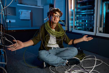 Foto de Ingeniero de redes de TI confuso mirando con incredulidad en cables Ethernet y cables ópticos en las manos. Dificultades en el trabajo - Imagen libre de derechos