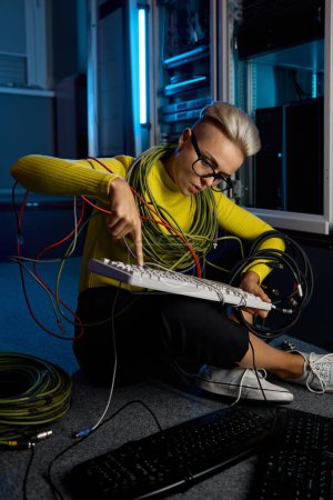 Téléchargez les photos : Jeune femme technicienne informatique enveloppée dans le fil de réparation clavier assis sur le sol de la salle des serveurs - en image libre de droit