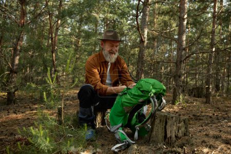 Téléchargez les photos : Voyageur sénior joyeux se préparant pour le repos après une randonnée en forêt. Incroyable aventure en forêt pour les retraités concept - en image libre de droit