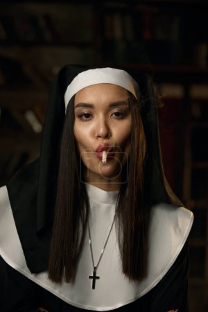 Foto de Retrato de monja sexy traviesa usando bata y tocado fumando cigarrillo. Concepto estropeado de religión - Imagen libre de derechos