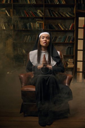 Foto de Joven mujer hermosa en vestido de monja fumar cigarrillo sentado en el sillón con las manos dobladas en posición de oración. Atractivo retrato santo pecador - Imagen libre de derechos