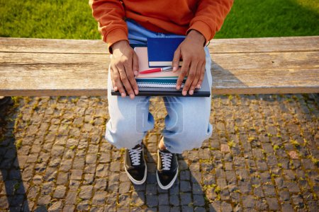 Photo for Student holding personal studying accessories as textbook, copy-book with pen and laptop while sitting on bench in university or college park - Royalty Free Image