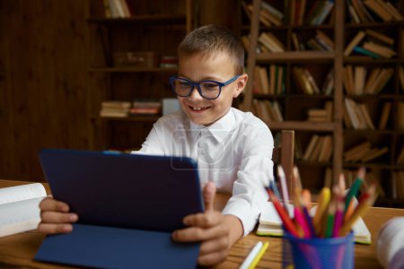 Photo for Cute smart boy schoolchild studying online using electronic device for watching funny video and learning while sitting at table, selective focus - Royalty Free Image