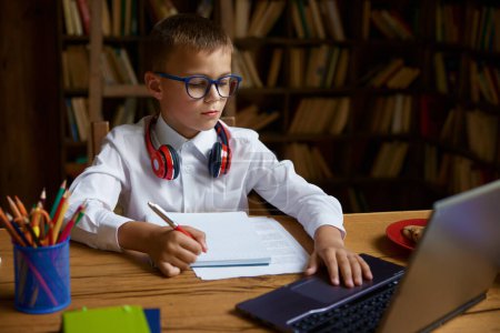 Téléchargez les photos : Petit garçon étudiant à la table à la maison assis devant un ordinateur portable. Concept d'éducation et de connaissance en ligne - en image libre de droit