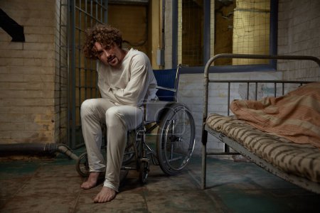 Téléchargez les photos : Un malade mental portant une camisole de force assise en fauteuil roulant au-dessus d'un hôpital psychiatrique. Homme fou souffrant de schizophrénie - en image libre de droit