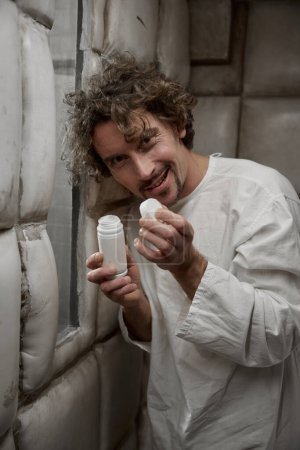 Foto de Hombre loco con trastorno psiquiátrico sonriendo ominosamente a la cámara sosteniendo abierto píldoras botella de pie en la habitación acolchada hospital - Imagen libre de derechos