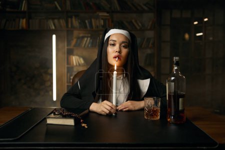 Téléchargez les photos : Une religieuse chrétienne immorale allume une cigarette en brûlant une bougie assise à table avec une bouteille de whisky ouverte. Portrait de femme vicieuse vêtue d'un costume monastique - en image libre de droit
