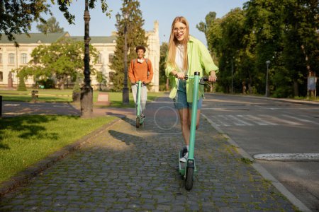 Téléchargez les photos : Compagnie Interraciale d'étudiants utilisant des véhicules électriques scooter équitation parmi le campus universitaire chemin - en image libre de droit