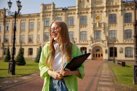 Téléchargez les photos : Portrait d'une adolescente sur fond de bâtiment universitaire. Confiant occasionnel jeune fille portant ordinateur portable à l'extérieur - en image libre de droit