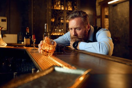 Photo for Depressed young adult man lost in sad thoughts drinking alcoholic beverage while sitting at bar counter - Royalty Free Image