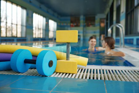 Photo for Aqua aerobics class, gymnastics training lesson at health club with swimming pool. Selective focus closeup on fitness sport equipment and instructor talking with client on blurred backdrop - Royalty Free Image