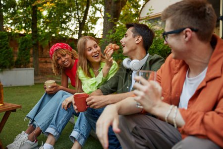 Foto de Diverso grupo de jóvenes organizando picnic fiesta al aire libre mientras acampan con furgoneta remolque. Vista de cerca de chicos hipster comer bocadillos dulces y beber té caliente - Imagen libre de derechos