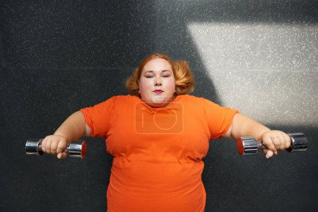 Téléchargez les photos : Femme en surpoids faisant de l'exercice efficace avec haltères poids couché sur le sol à la salle de gym vue aérienne - en image libre de droit