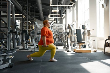 Foto de Mujer obesa confiada haciendo embestidas hacia adelante con pesas mientras entrena en el club deportivo moderno gimnasio - Imagen libre de derechos