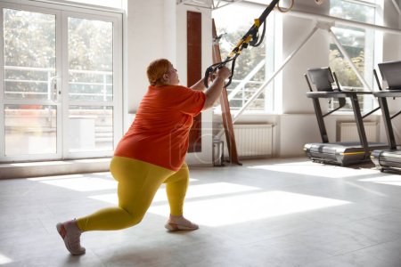 Téléchargez les photos : Femme joufflu travaillant dans un programme d'entraînement de poids corporel en utilisant des boucles trx. Exercices de résistance totale pour le poids perdu - en image libre de droit