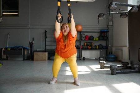 Téléchargez les photos : Femme joufflu travaillant dans un programme d'entraînement de poids corporel en utilisant des boucles trx. Exercices de résistance totale pour le poids perdu - en image libre de droit