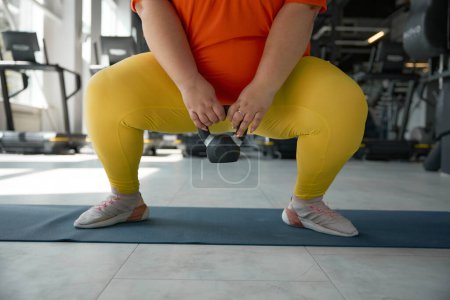 Téléchargez les photos : Vue de la culture de la femme obèse en surpoids accroupi avec kettlebell pendant l'entraînement à la salle de gym. Loisirs sportifs pour perdre du poids et concept de soins de santé - en image libre de droit