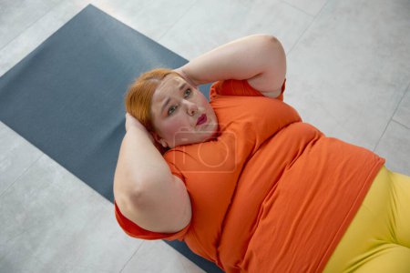 Téléchargez les photos : Femme en surpoids faisant abdos croquer couché sur le tapis. Fille obèse ayant séance d'entraînement physique sur l'entraînement personnel à la salle de gym - en image libre de droit
