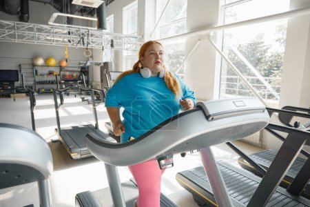 Téléchargez les photos : Femme obèse active courir sur la piste de tapis roulant en profitant de l'exercice cardio pour brûler les calories. Défi sportif pour mincir - en image libre de droit