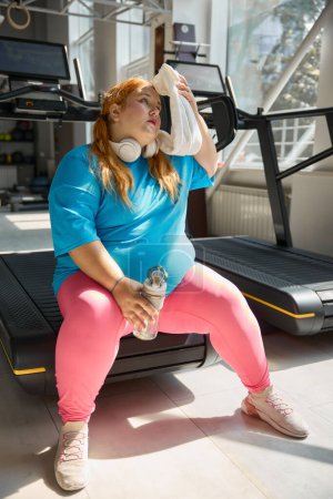 Photo for Tired plus size woman wiping sweat from her face with towel feeling exhaustion after cardio training on treadmill machine - Royalty Free Image