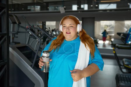 Photo for Portrait of young overweight woman wearing sportive clothes and headphones holding bottle of water standing over gym. Teenage girl ready for training on treadmill machine - Royalty Free Image