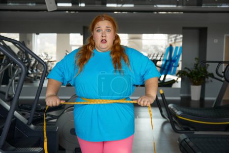 Foto de Retrato mujer linda con exceso de peso ata a sí misma con cinta métrica de pie en el gimnasio. Dieta para adelgazar y entrenamiento deportivo para mujeres obesas - Imagen libre de derechos