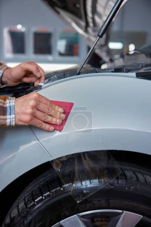 Photo for Closeup master hand with rubber scraper applying sticking protective film on car at vehicle workshop service station - Royalty Free Image