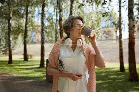 Téléchargez les photos : Joyeux retraité portant des écouteurs et tenue à la mode boire du café à travers le parc de la ville. Plaisir estival pendant la retraite - en image libre de droit