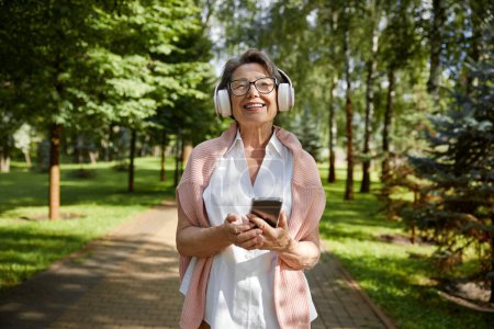 Téléchargez les photos : Portrait de vieille femme satisfaite utilisant mobile profiter de la journée d'été tout en marchant dans le parc public - en image libre de droit