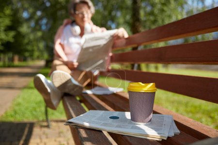 Photo for Focus on coffee cup and newspaper and restful senior woman reading articles in daily news on park bench on blurred background - Royalty Free Image
