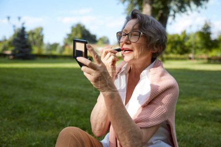 Téléchargez les photos : Femme âgée appliquant rouge à lèvres faisant maquillage pour sourire charmant dans un miroir compact tout en se reposant en plein air dans le parc de la ville - en image libre de droit