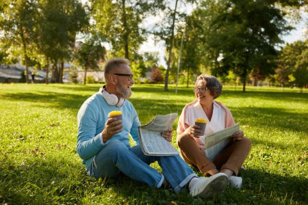 Photo for Senior loving couple drinking coffee and reading newspaper while relaxing on green lawn grass outdoors in park - Royalty Free Image
