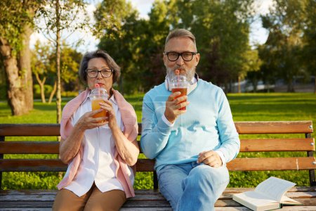 Téléchargez les photos : Portrait de couple âgé détendu buvant un cocktail rafraîchissant dans une tasse à emporter assis sur un banc dans un parc. Bonne vie à la retraite - en image libre de droit