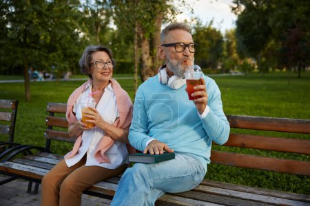 Téléchargez les photos : Portrait de couples âgés détendus buvant des cocktails rafraîchissants dans une tasse à emporter assis sur un banc dans un parc. Bonne vie à la retraite - en image libre de droit