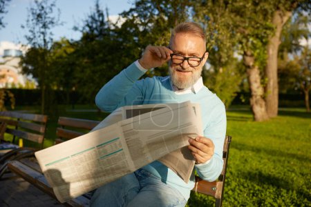 Photo for Confident bearded mature man reading daily newspaper on park bench. Morning pastime on retirement - Royalty Free Image