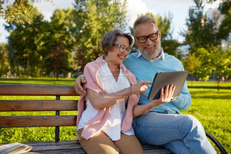 Téléchargez les photos : Heureux couple de personnes âgées gaies en utilisant une tablette numérique tout en se reposant sur le banc du parc. Concept moderne de mode de vie des personnes âgées - en image libre de droit