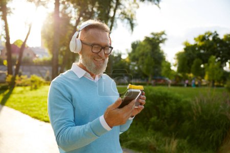 Photo for Happy casual senior man wearing headset having video conference or watching funny movie on mobile drinking coffee on walk in public park - Royalty Free Image