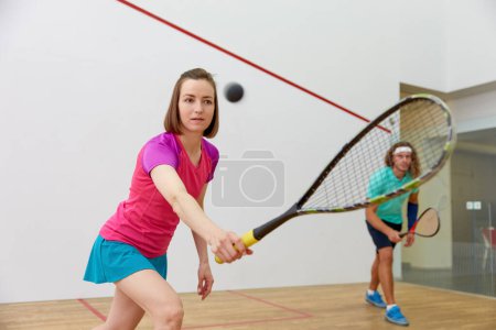 Photo for Young sportive couple practicing squash at indoor court. Focus on attractive woman hitting ball with racket - Royalty Free Image