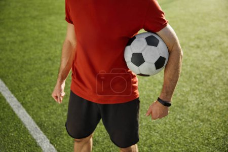Photo for Crop view of football trainer or soccer player holding ball standing over green city stadium background. Good sportive day, successful training and recreational pursuit - Royalty Free Image