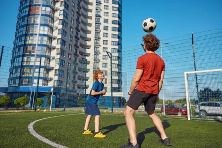 Photo for Dad showing trick to his son hitting soccer ball by head. Excited family training at football field during summer vacation - Royalty Free Image