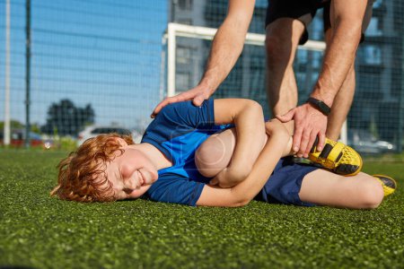 Photo for Closeup of boy child hurts his leg feeling pain during training football match. Supportive father trying to help son. Sportive injury and first aid - Royalty Free Image