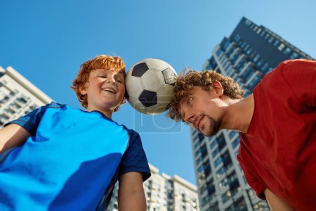 Téléchargez les photos : Gros plan de l'homme adulte et garçon enfant tenant ballon de football entre le front vue du bas sur le fond du gratte-ciel de la ville. Compétition familiale, combat fils contre père - en image libre de droit