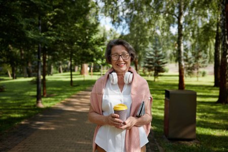 Photo for Happy retired woman wearing headphones and trendy outfit with coffee walking through city park. Summer enjoyment during retirement - Royalty Free Image