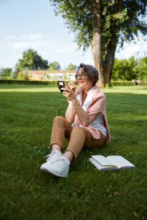 Photo for Senior woman applying lipstick doing makeup for charming smile in compact mirror while resting outdoor in city park on the lawn - Royalty Free Image