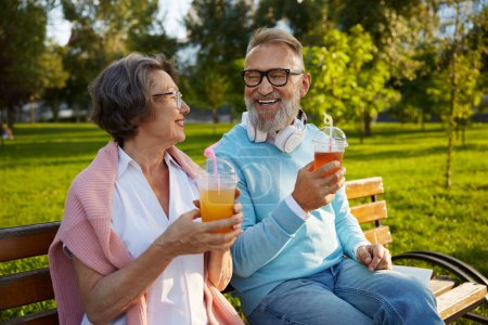 Téléchargez les photos : Portrait de couple âgé détendu buvant un cocktail rafraîchissant dans une tasse à emporter assis sur un banc dans un parc. Bonne vie à la retraite - en image libre de droit