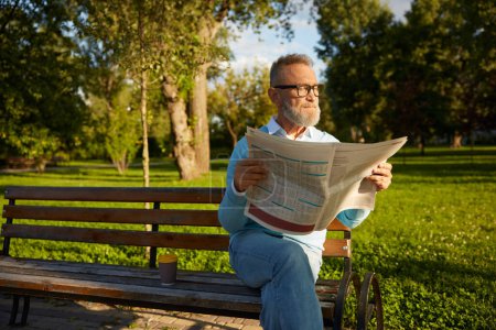 Foto de Hombre maduro barbudo seguro leyendo diario en el banco del parque. Pasatiempo matutino de jubilación - Imagen libre de derechos